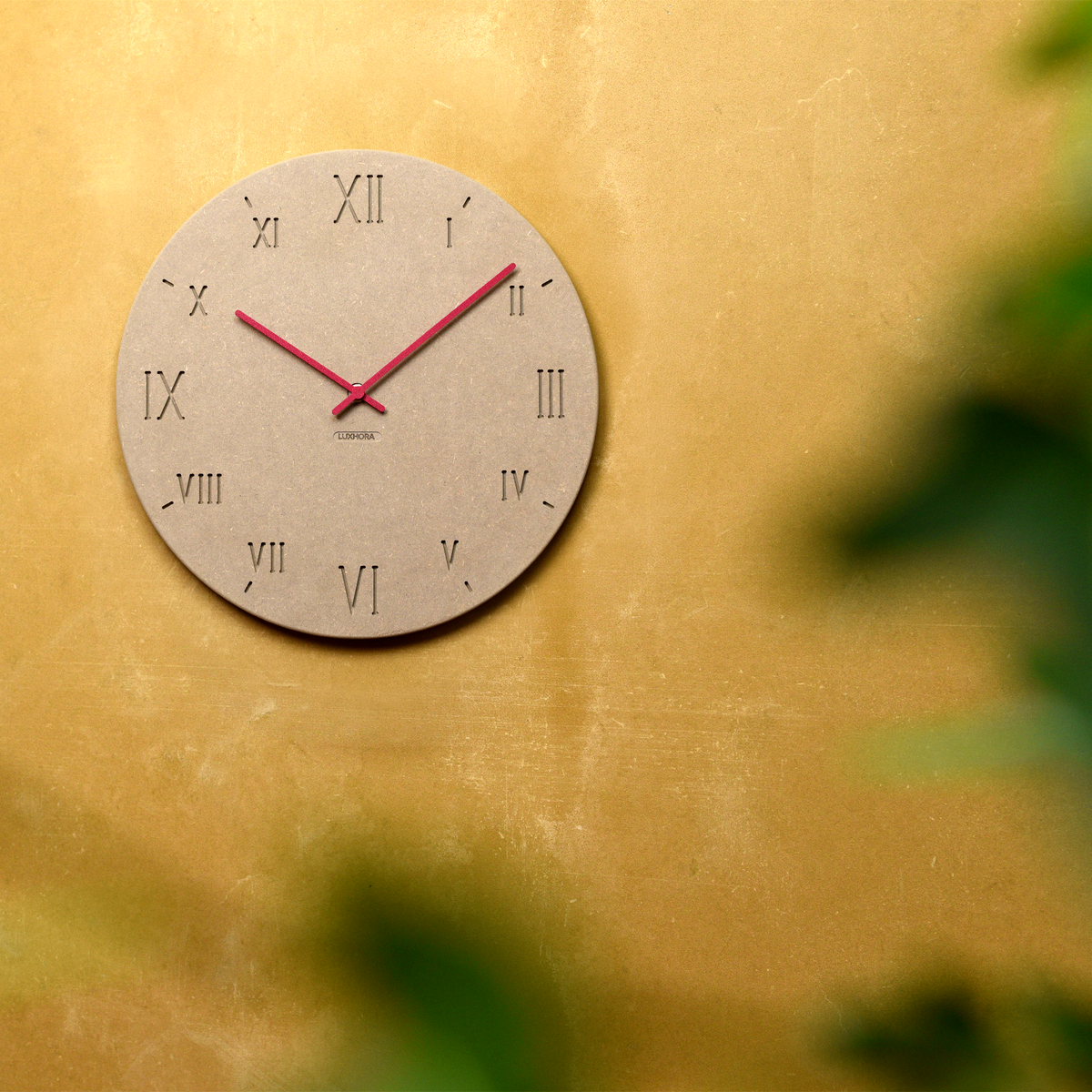 LUXHORA Romana Wanduhr in Valchromat Khaki an einer Wand, Pflanze im Vordergrund