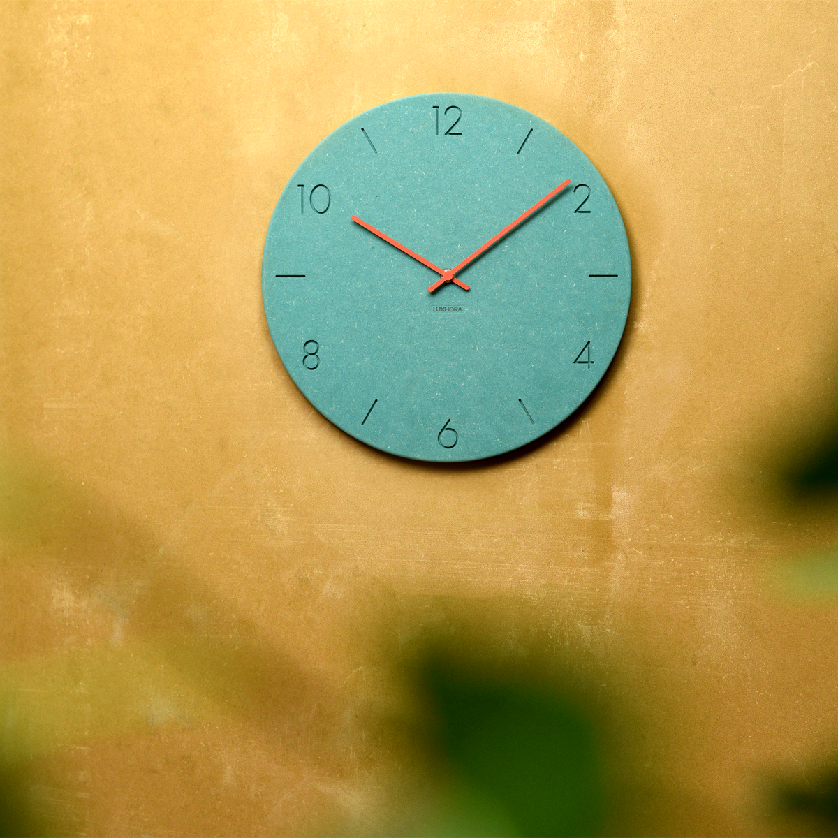 LUXHORA Sena Wanduhr in Valchromat Minzgrün an einer Wand, Pflanze im Vordergrund